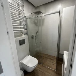 DONG BAU GROUP SPÓŁKA Z OGRANICZONĄ ODPOWIEDZIALNOŚCIĄ - Nowoczesna łazienka po remoncie: kabina prysznicowa ze szklanymi drzwiami, podwieszana toaleta, grzejnik drabinkowy i drewnopodobne płytki podłogowe. Minimalistyczny design.