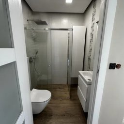 DONG BAU GROUP SPÓŁKA Z OGRANICZONĄ ODPOWIEDZIALNOŚCIĄ - Nowoczesna łazienka z kabiną prysznicową typu walk-in, drewnopodobną podłogą i białymi meblami, widziana zza uchylonych drzwi. Minimalistyczny design.