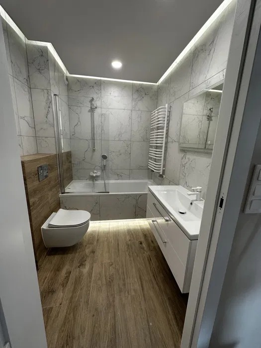 Nowoczesna łazienka z marmurowymi płytkami, drewnopodobną podłogą i podświetleniem LED. Umywalka z szafką, toaleta podwieszana i wanna z parawanem.