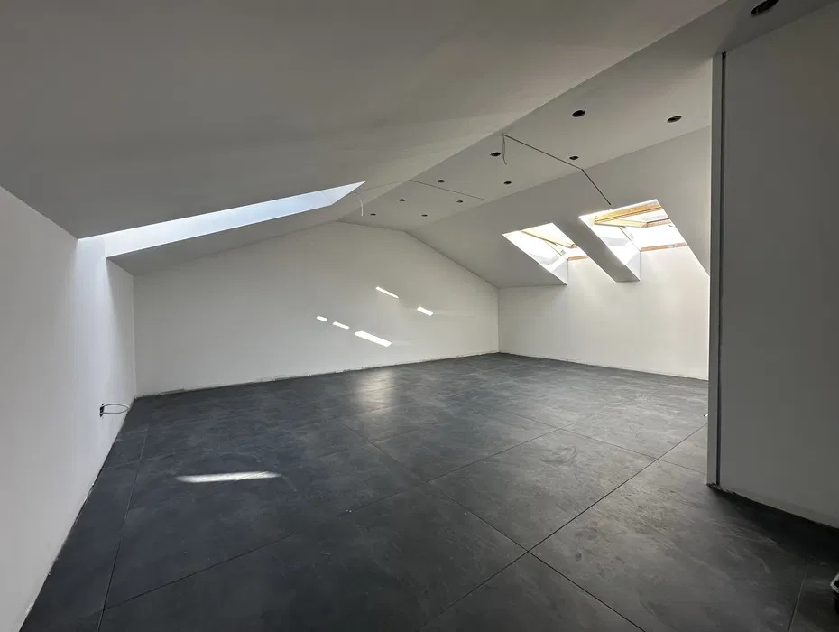 Poddasze po remoncie: białe ściany, ciemna podłoga z płytek, dwa okna dachowe wpuszczające światło. Widoczne przygotowania do montażu oświetlenia.