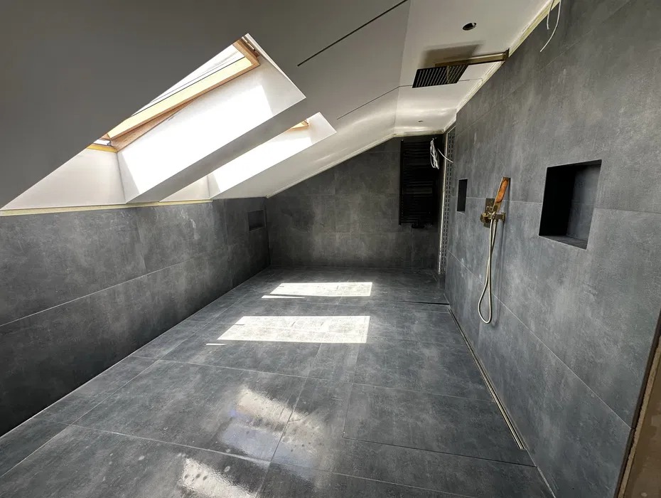 Szara łazienka na poddaszu z oknami dachowymi, wykończona płytkami. Widoczna armatura prysznicowa i grzejnik. Minimalistyczny design z wnękami w ścianie.