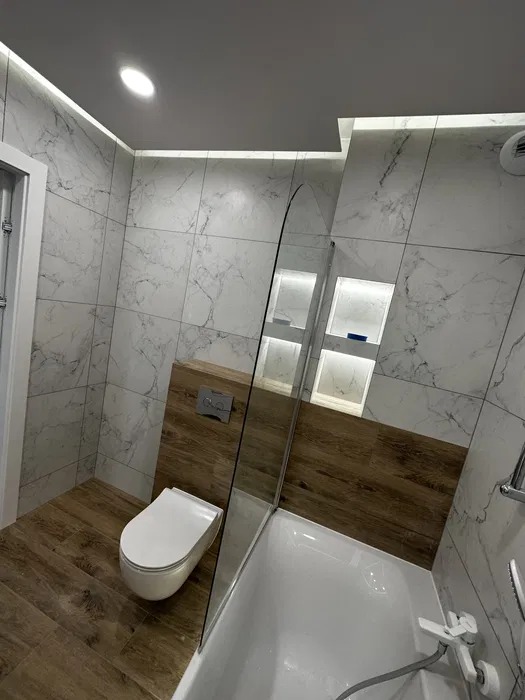 Nowoczesna łazienka po remoncie: białe marmurowe płytki, drewnopodobna podłoga, podświetlane wnęki, minimalistyczna toaleta i szklany parawan nawannowy.