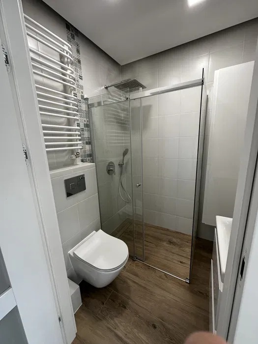 Nowoczesna łazienka po remoncie: kabina prysznicowa ze szklanymi drzwiami, podwieszana toaleta, grzejnik drabinkowy i drewnopodobne płytki podłogowe. Minimalistyczny design.