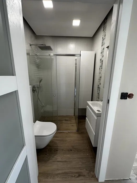 Nowoczesna łazienka z kabiną prysznicową typu walk-in, drewnopodobną podłogą i białymi meblami, widziana zza uchylonych drzwi. Minimalistyczny design.