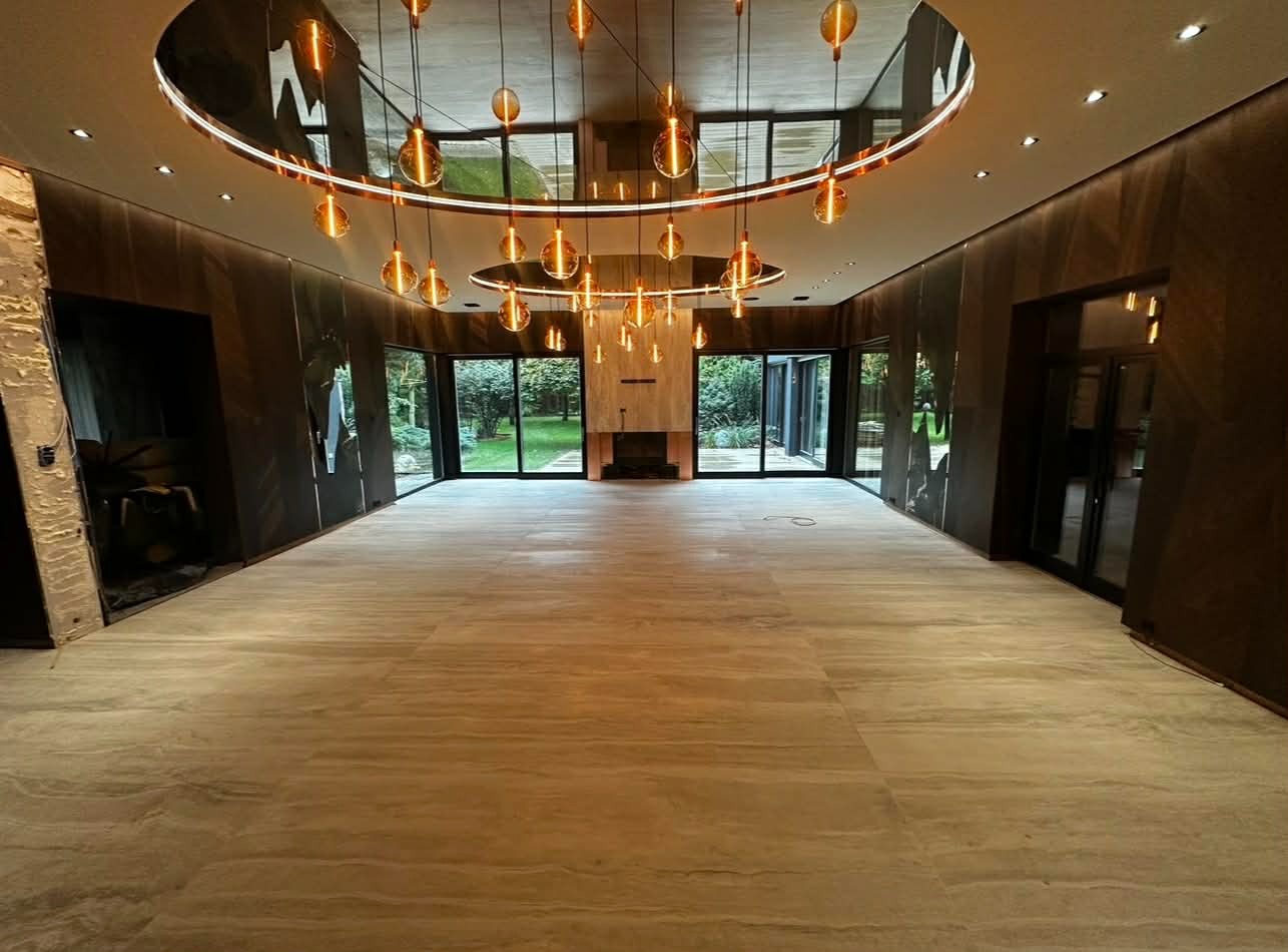 Przestronne wnętrze z jasną podłogą, ciemnymi panelami ściennymi i okrągłą lampą sufitową z wiszącymi, bursztynowymi kulami. Widok na ogród przez duże okna.