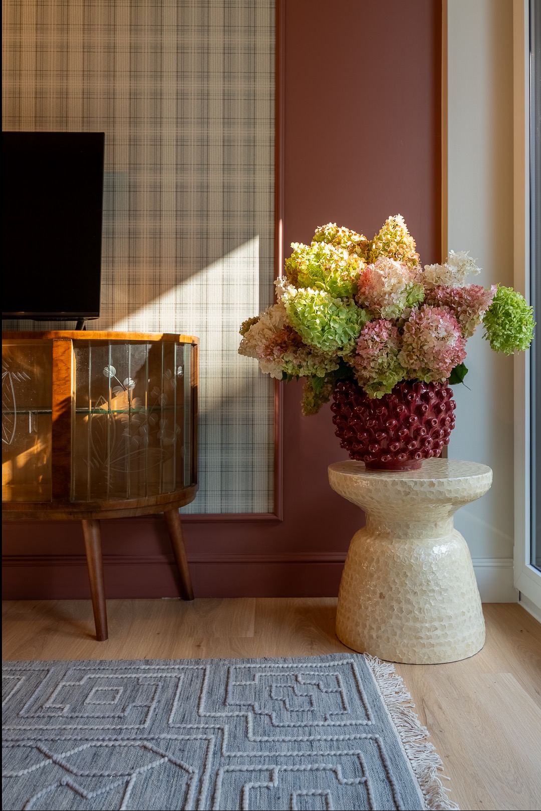 Stylowe wykończenie wnętrza w Raszynie: dekoracyjna komoda, wzorzysta tapeta i bukiet hortensji w bordowym wazonie na ceramicznym stoliku. Dywan z geometrycznym wzorem.