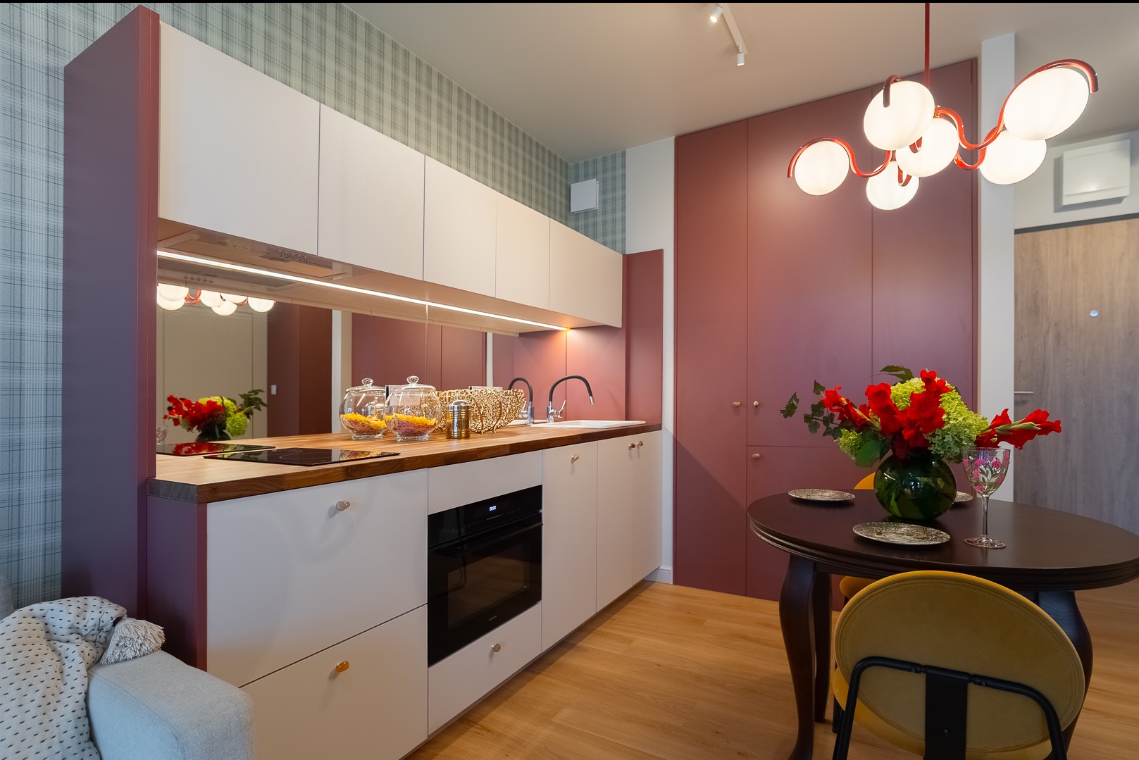 Nowoczesna kuchnia z białymi szafkami, drewnianym blatem i lustrem nad zlewem. Czerwone akcenty kolorystyczne i designerska lampa. Widok na stół z kwiatami.