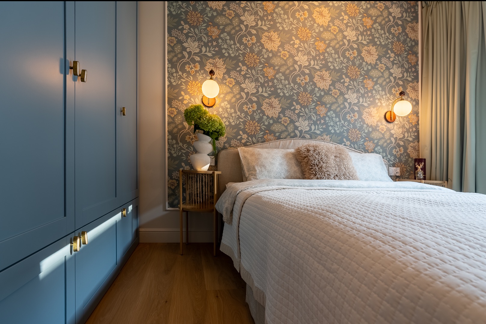Stylowa sypialnia z niebieską szafą wnękową, wzorzystą tapetą i lampkami nocnymi. Widoczny fragment łóżka z dekoracyjną poduszką i drewniana podłoga.