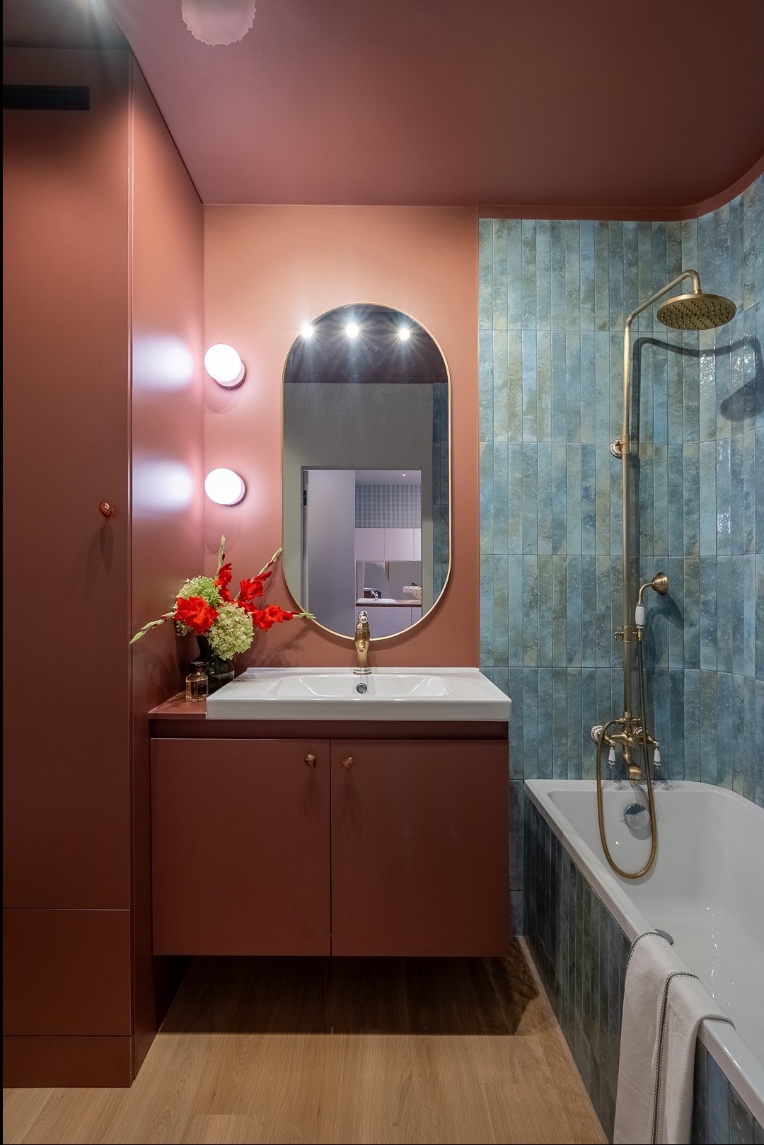 Nowoczesna łazienka w odcieniach terakoty i błękitu. Umywalka z szafką, owalne lustro z oświetleniem, bateria w kolorze złota, błękitne płytki na ścianie przy wannie.
