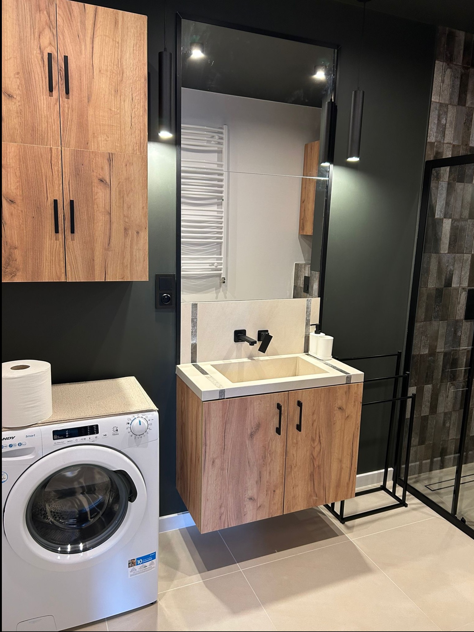 Nowoczesna łazienka po remoncie: szafki z drewnianym wzorem, umywalka, pralka Candy, czarne lampy wiszące i kabina prysznicowa z ciemnymi płytkami.