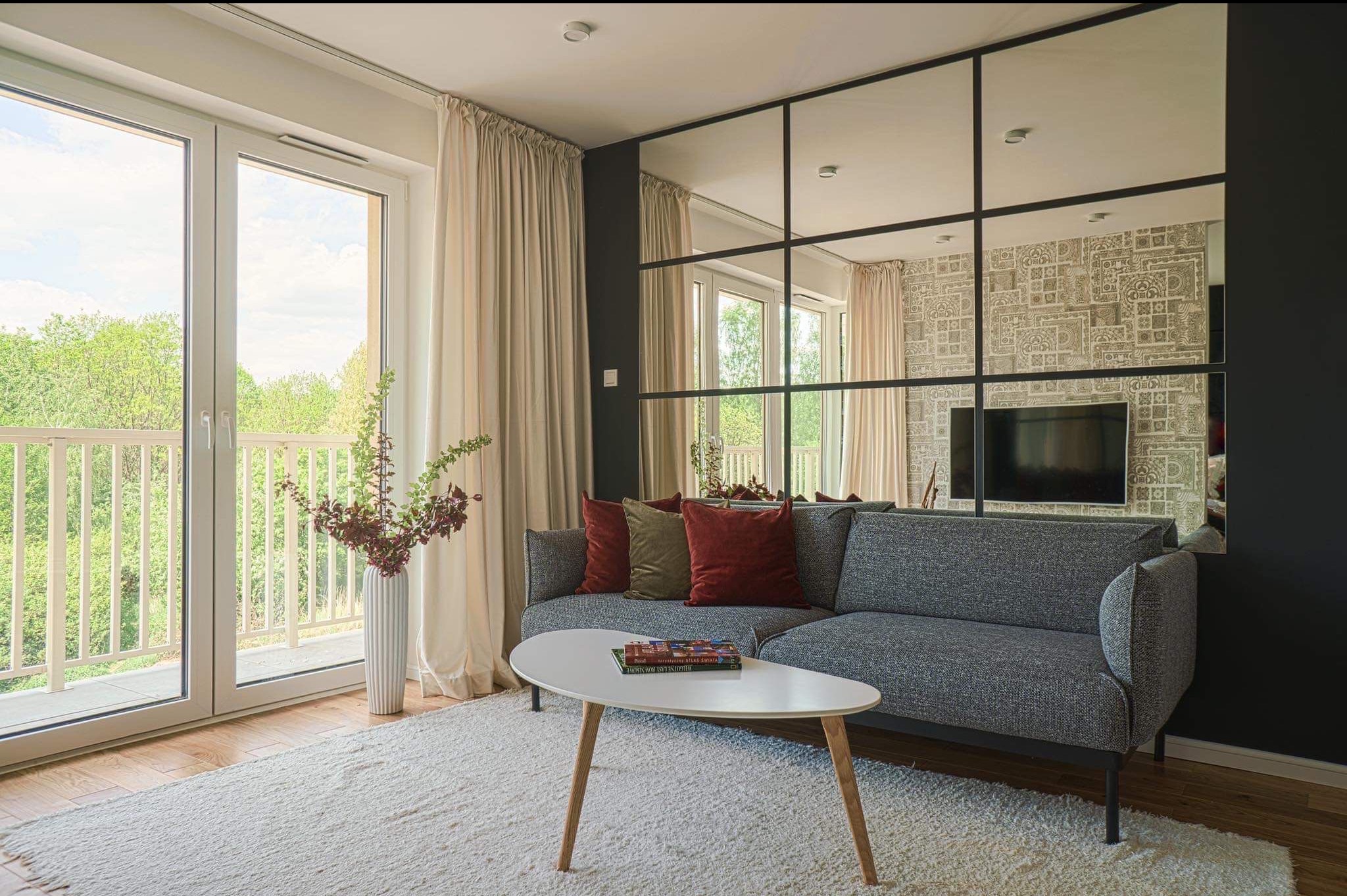 Jasny salon z balkonem: szara sofa z poduszkami, biały dywan, stolik kawowy. Lustro w czarnej ramie odbija wzorzystą tapetę i TV. Widok na zieleń z balkonu.