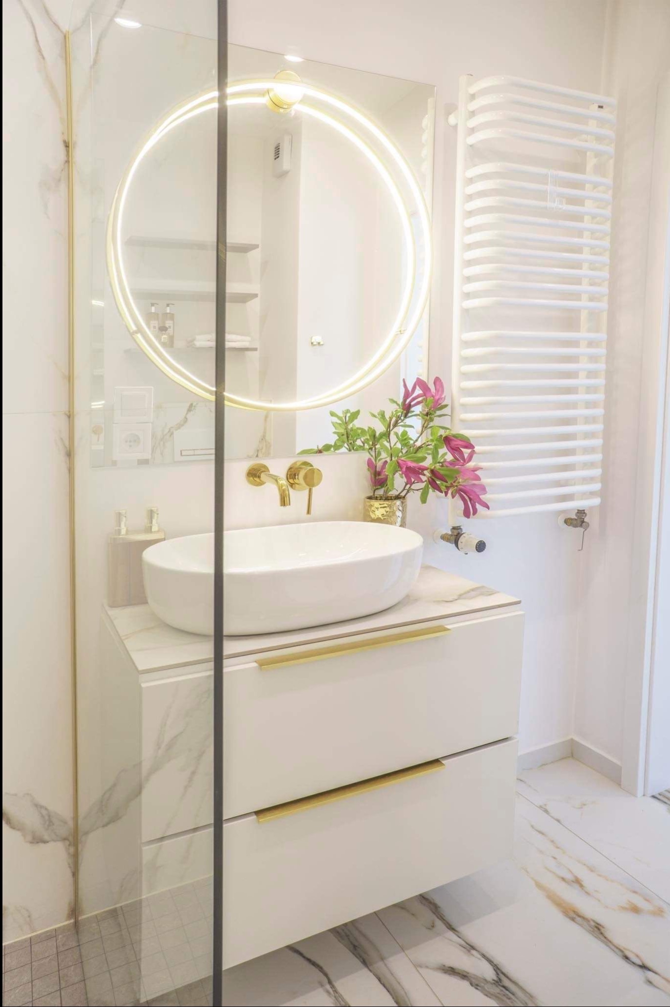 Elegancka łazienka z okrągłym lustrem LED, białą umywalką nablatową, złotą armaturą i grzejnikiem drabinkowym. Marmurowe płytki na ścianach i podłodze.