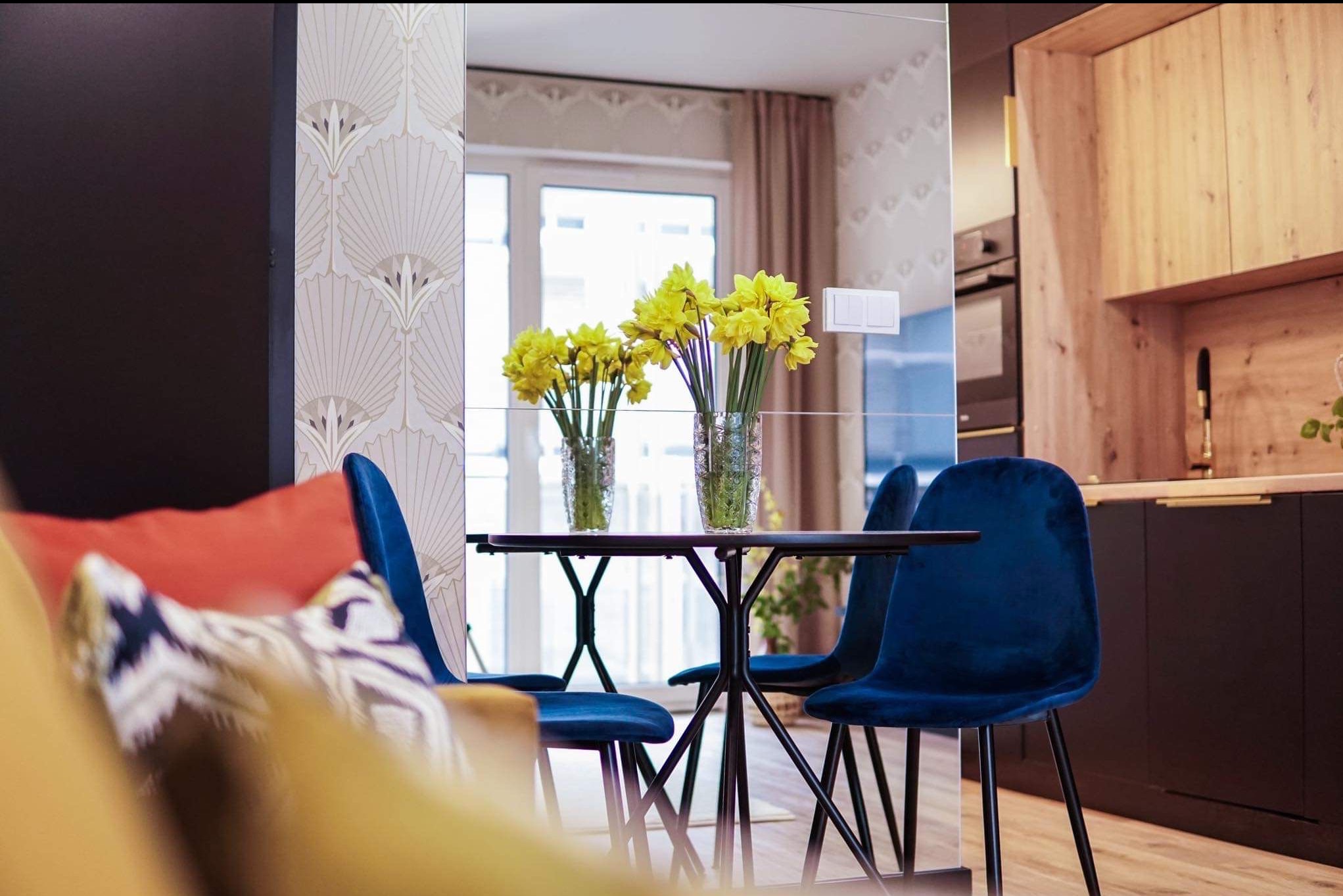 Stylowe wnętrze: ciemne meble kuchenne z drewnianymi akcentami, stół z bukietem żółtych kwiatów, welurowe krzesła w odcieniach granatu i wzorzysta tapeta w tle.