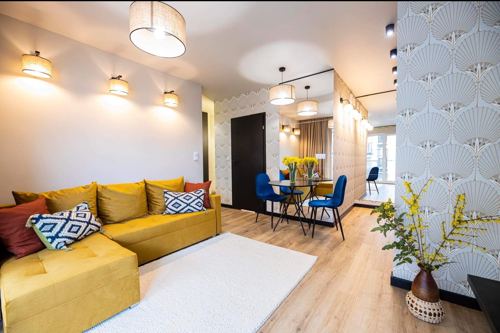 Salon z żółtą sofą narożną, okrągłym stołem i wzorzystą tapetą w stylu art deco. Oświetlenie punktowe i dywan dopełniają aranżację.