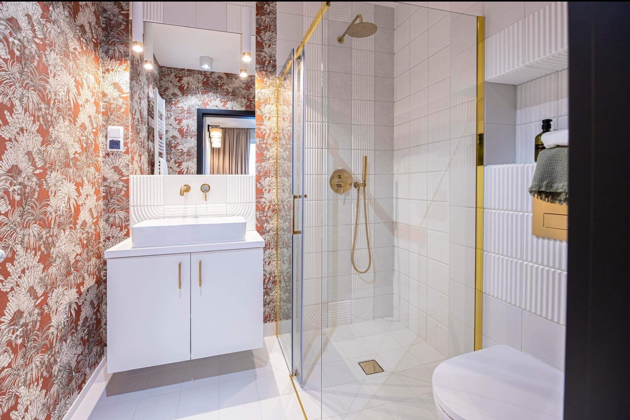 Nowoczesna łazienka z kabiną prysznicową, białą umywalką na szafce i wzorzystą tapetą w odcieniach brązu. Złote akcenty armatury i wykończeń dodają elegancji.