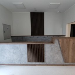 DECHA - Nowoczesna recepcja z drewnianymi i betonowymi elementami. Widoczne szafki z szufladami, drewniane lamele i oświetlenie sufitowe. Minimalistyczny design.