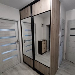 DECHA - Szafa przesuwna z lustrami i drewnianym wykończeniem, czarne obramowanie, widok z perspektywy podłogi, w tle drzwi i komoda, jasne wnętrze.