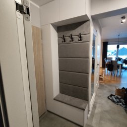 DECHA - Biała szafa wnękowa z siedziskiem i panelem w odcieniach szarości, z trzema czarnymi haczykami na ubrania. Widok na wnętrze domu przez otwarte drzwi.