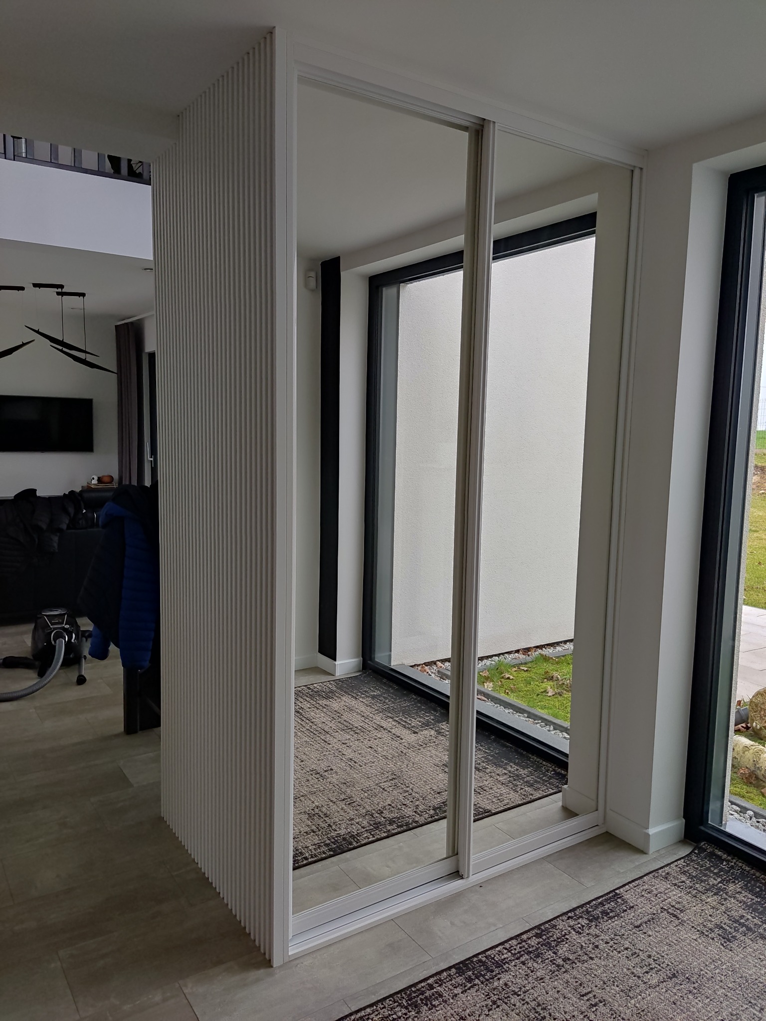 Biała szafa przesuwna z lustrami, oddzielająca przestrzeń. Widoczne ryflowane panele boczne i fragment salonu z sofą. Nowoczesny design, jasne wnętrze.