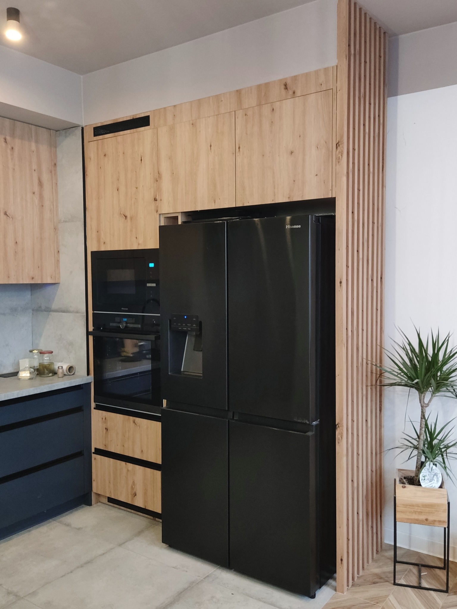 Nowoczesna kuchnia z drewnianymi frontami, czarną lodówką side-by-side i zabudowaną piekarnikiem. Dekoracyjny panel z listewek po prawej stronie.