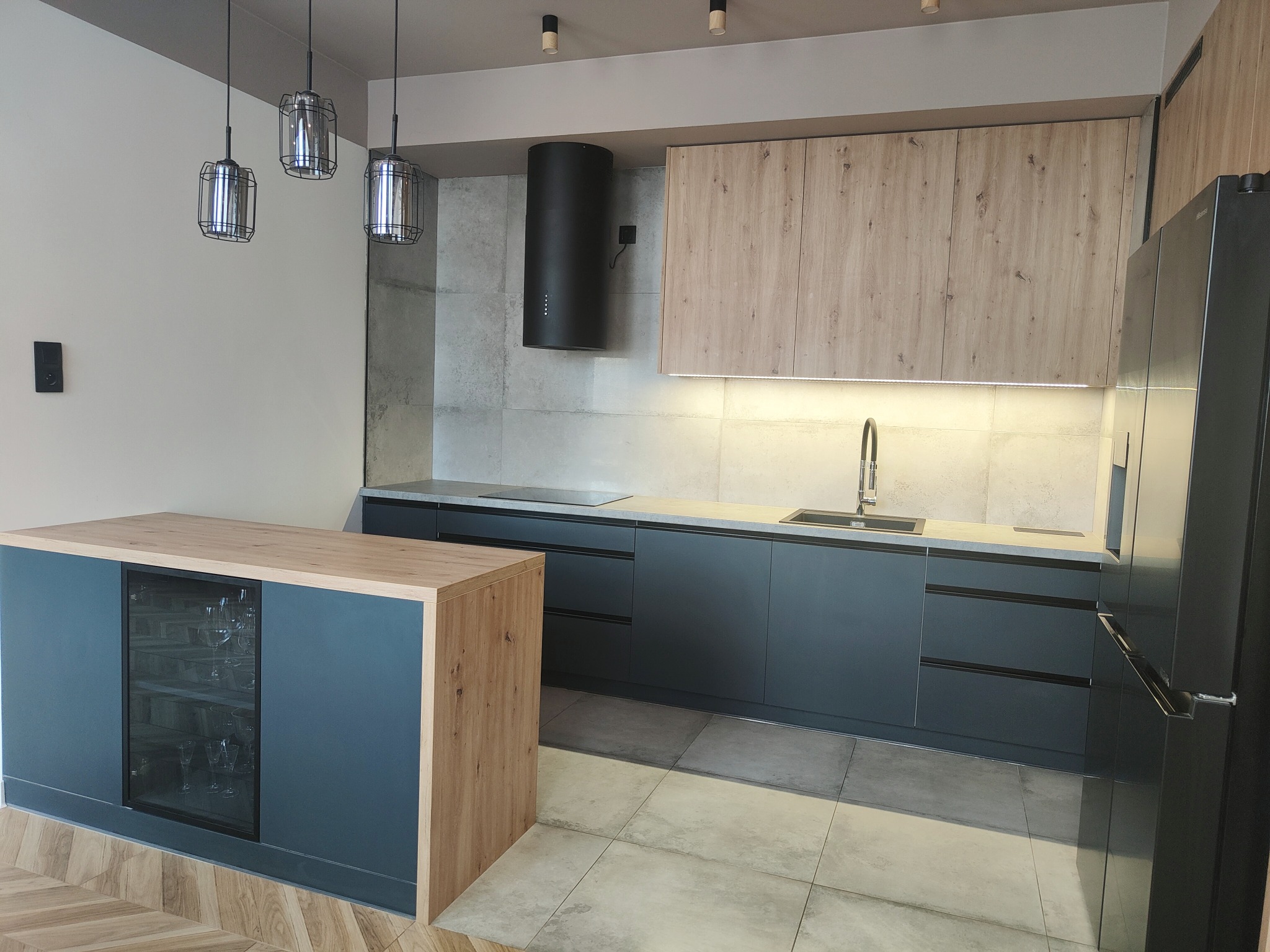 Nowoczesna kuchnia z szarymi szafkami i drewnianymi akcentami, wyspą z chłodziarką na wino, oświetleniem punktowym i wiszącymi lampami w metalowych klatkach.