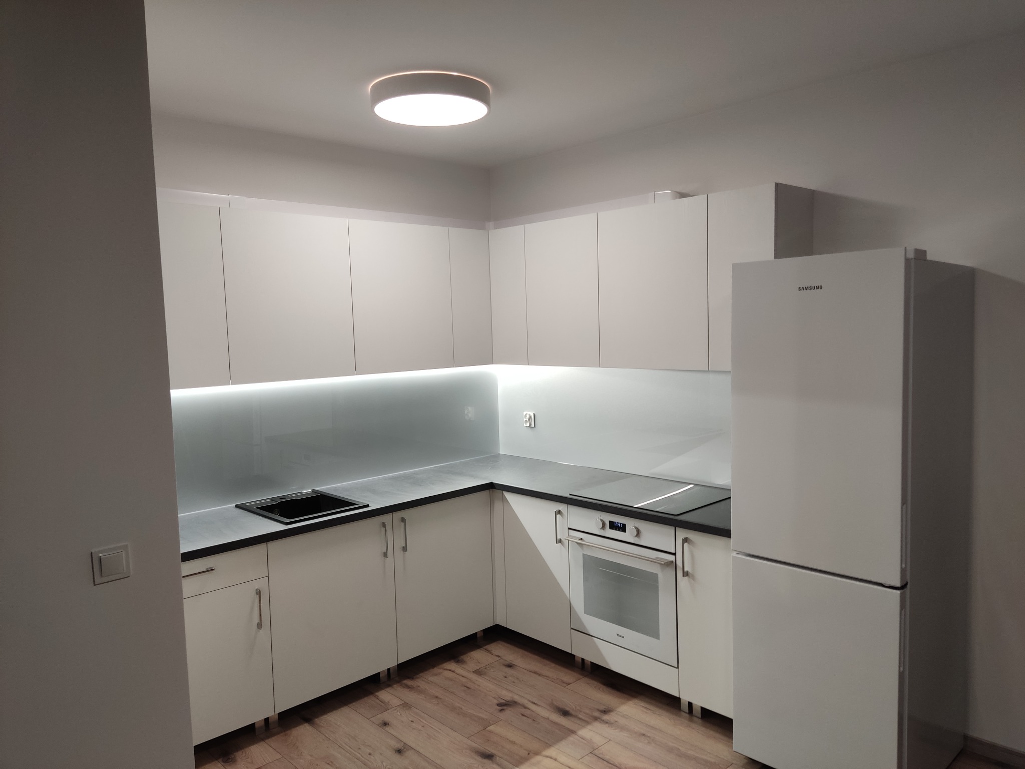 Minimalistyczna, jasna kuchnia z białymi frontami, szarym blatem i podświetleniem LED. Widoczny zlew, płyta indukcyjna i lodówka Samsung. Parkiet na podłodze.