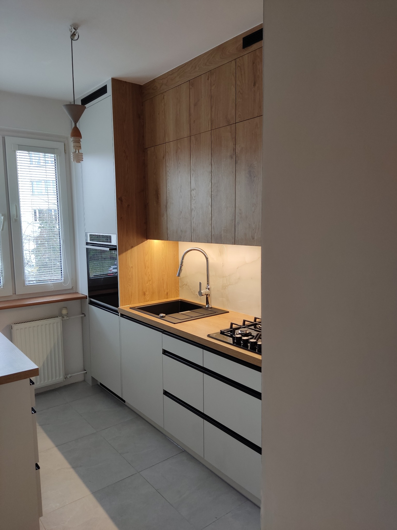 Nowoczesna kuchnia z drewnianymi szafkami górnymi i białymi dolnymi, czarnymi akcentami. Uwagę przykuwa oświetlenie LED pod szafkami i minimalistyczny design.