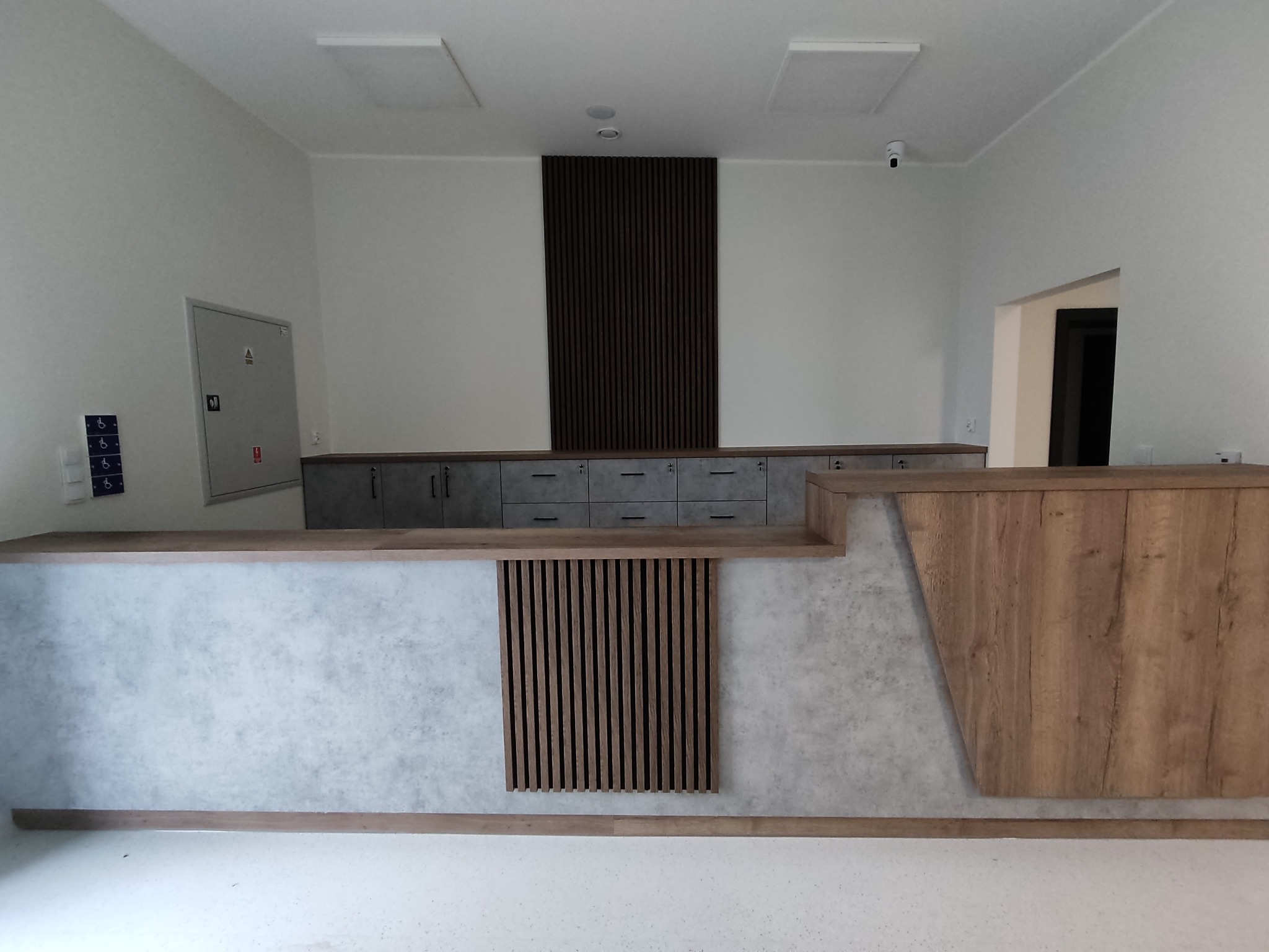 Nowoczesna recepcja z drewnianymi i betonowymi elementami. Widoczne szafki z szufladami, drewniane lamele i oświetlenie sufitowe. Minimalistyczny design.
