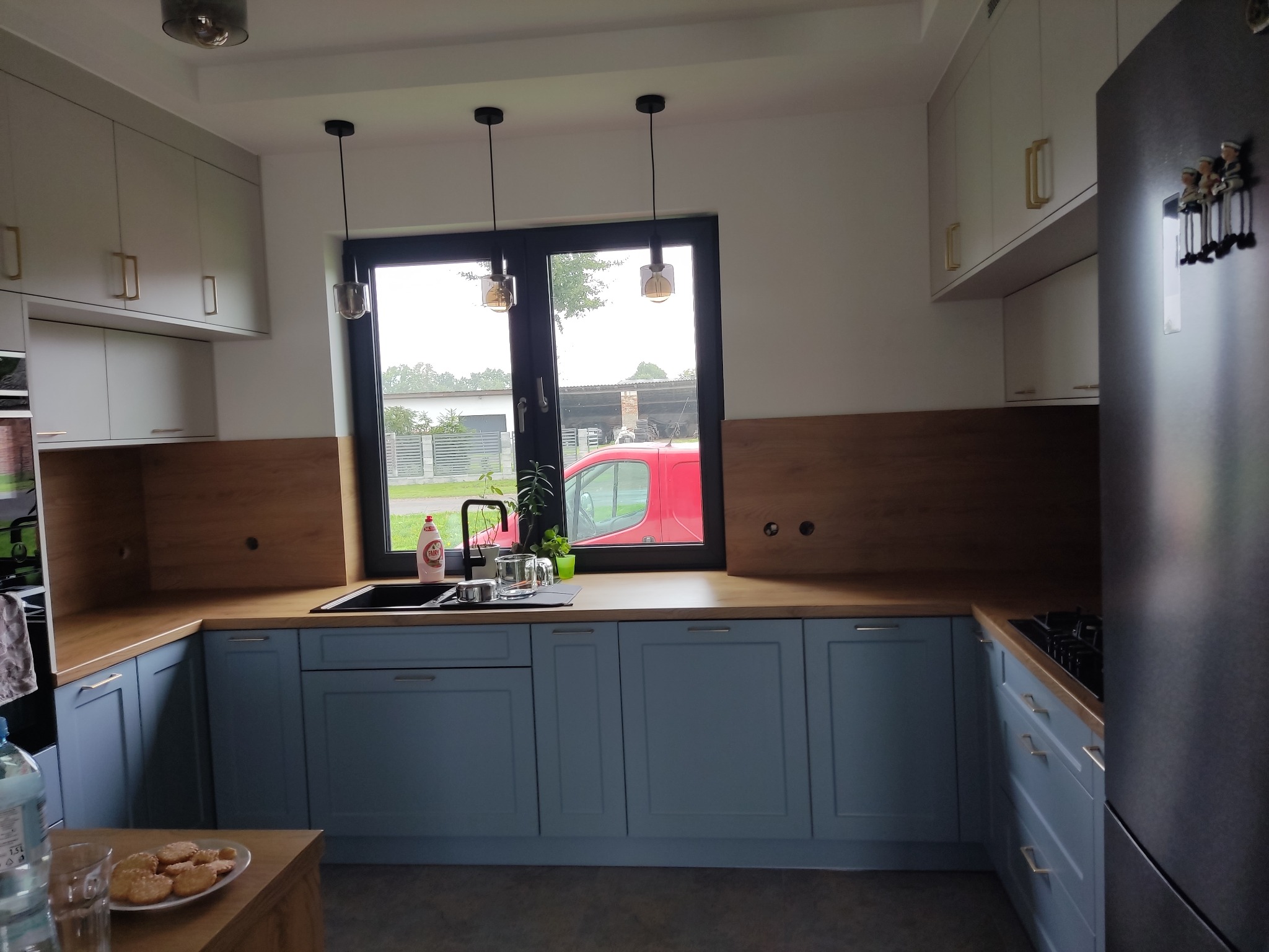 Jasna kuchnia z błękitnymi szafkami i drewnianym blatem, oświetlona trzema lampami wiszącymi. Za oknem widoczny czerwony samochód i zieleń.