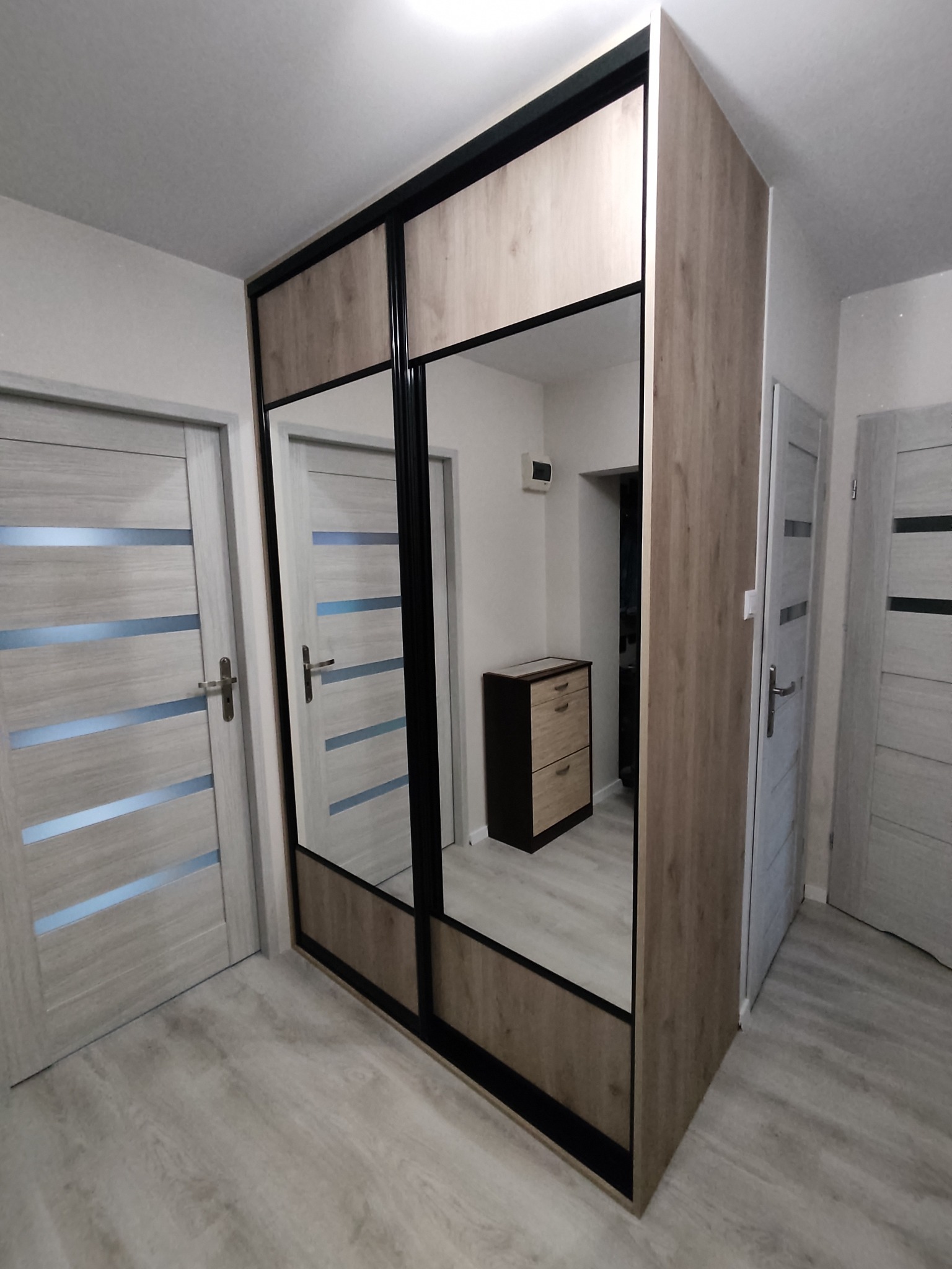 Szafa przesuwna z lustrami i drewnianym wykończeniem, czarne obramowanie, widok z perspektywy podłogi, w tle drzwi i komoda, jasne wnętrze.