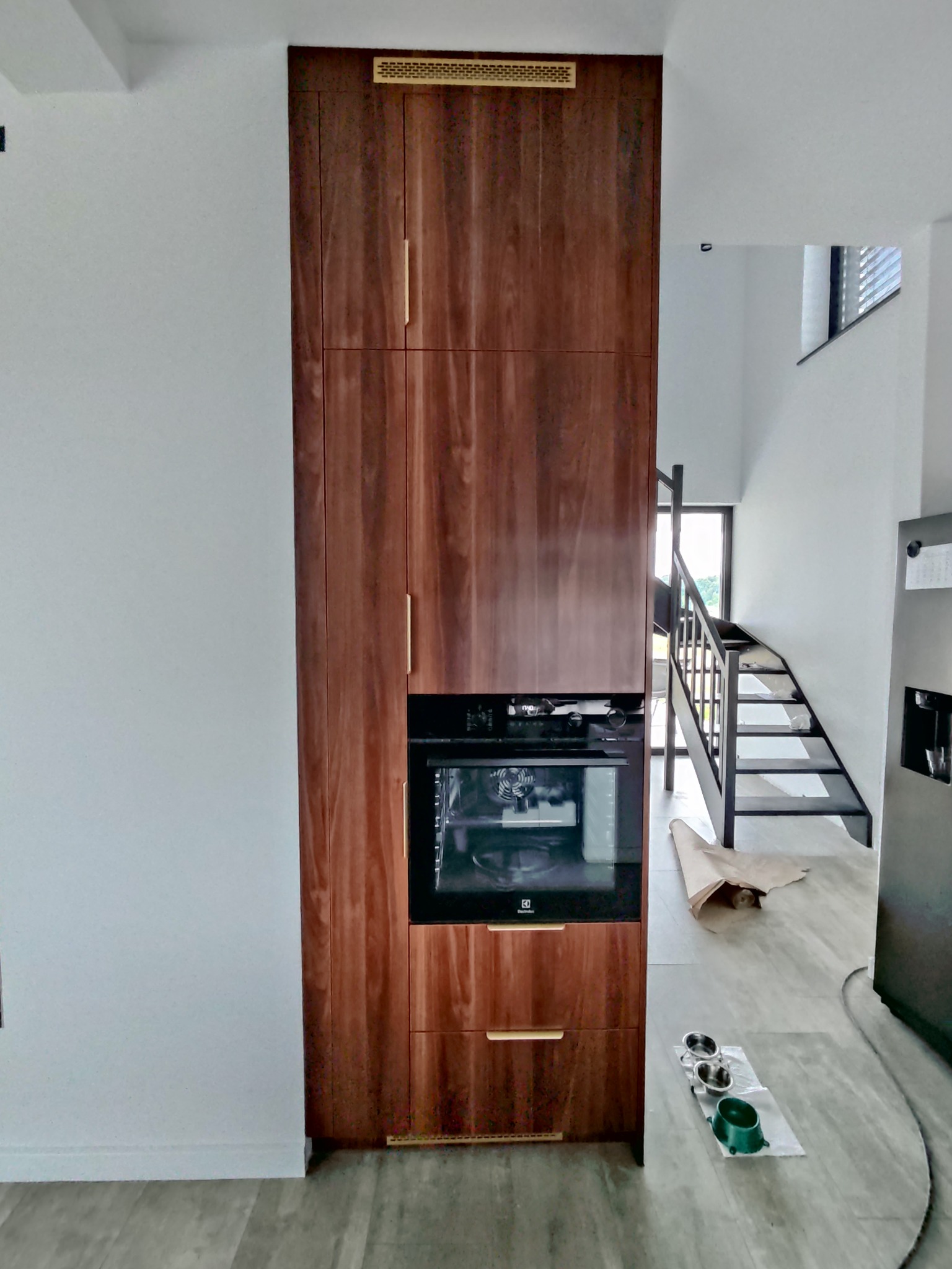 Wysoka, drewniana zabudowa kuchenna z piekarnikiem i złotymi uchwytami, w nowoczesnym wnętrzu z metalowymi schodami i szarą podłogą. Widoczne miski dla zwierząt.