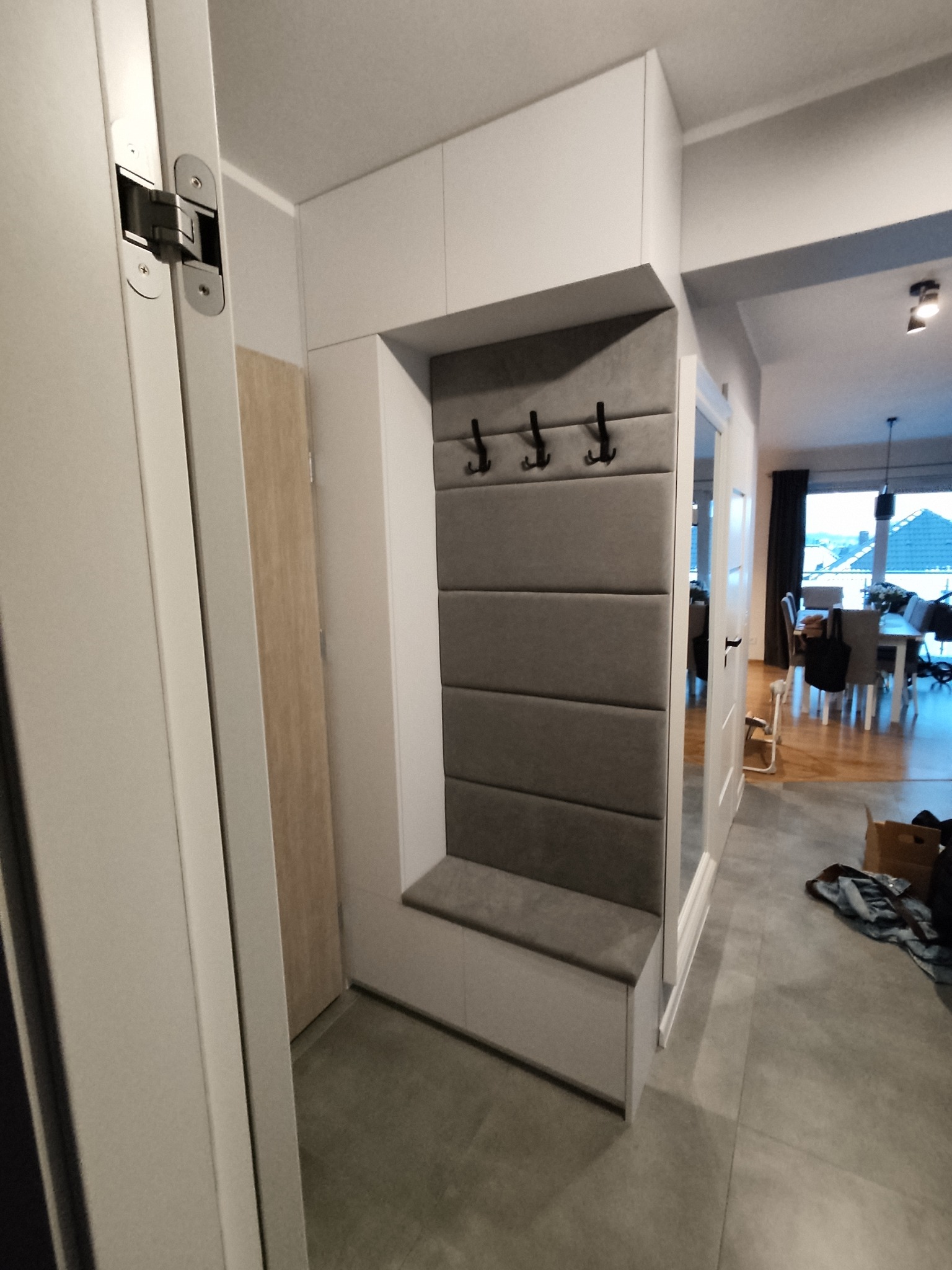 Biała szafa wnękowa z siedziskiem i panelem w odcieniach szarości, z trzema czarnymi haczykami na ubrania. Widok na wnętrze domu przez otwarte drzwi.
