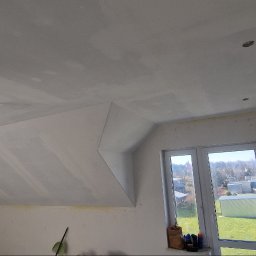 Patryk Sobejko Dom w Detalu - Poddasze po szpachlowaniu, widoczne łączenia płyt gipsowych na skosach i suficie, przygotowane do dalszych prac wykończeniowych. Okno z widokiem na zewnątrz.