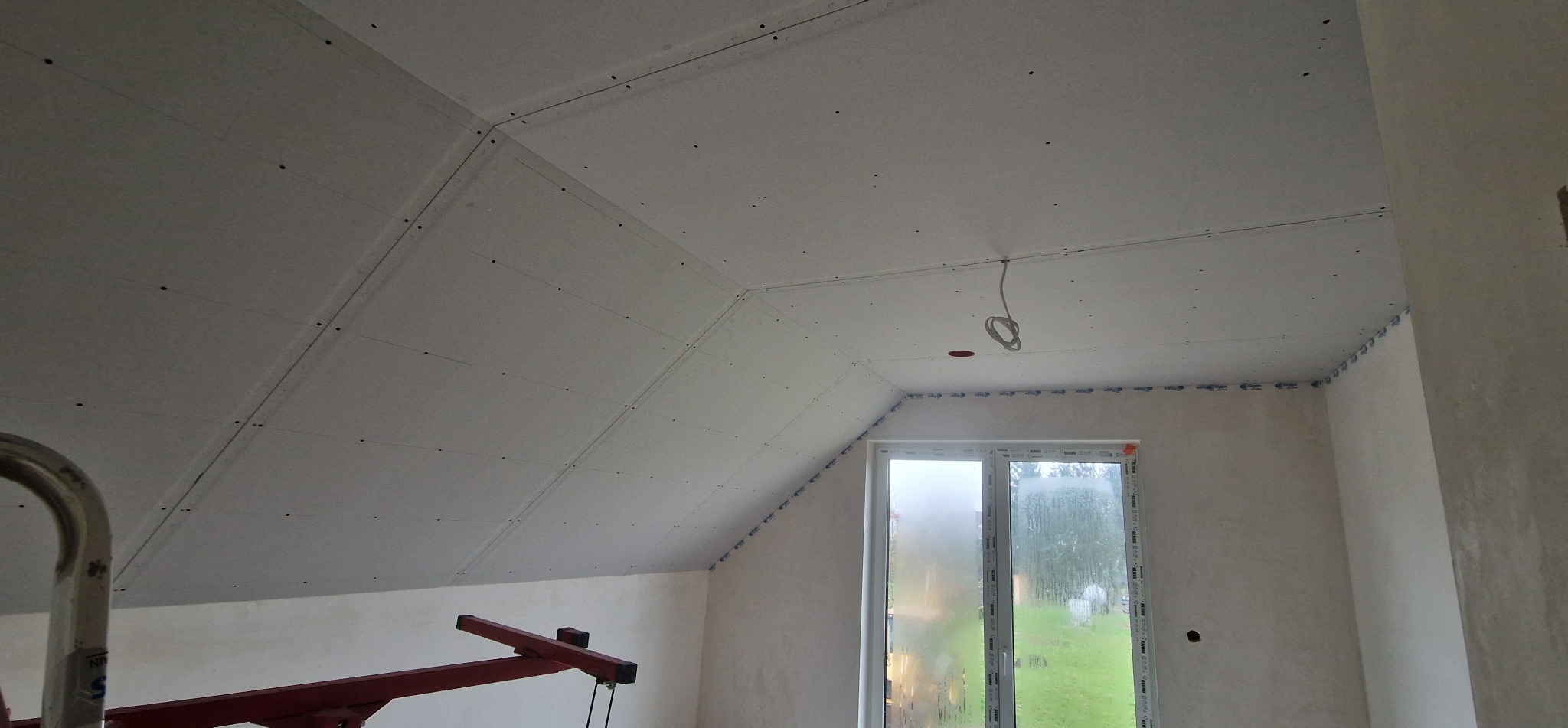 Skośny sufit poddasza z zamontowanymi płytami gipsowo-kartonowymi, widoczne łączenia i otwór na oświetlenie. Okno w tle. Etap prac wykończeniowych.