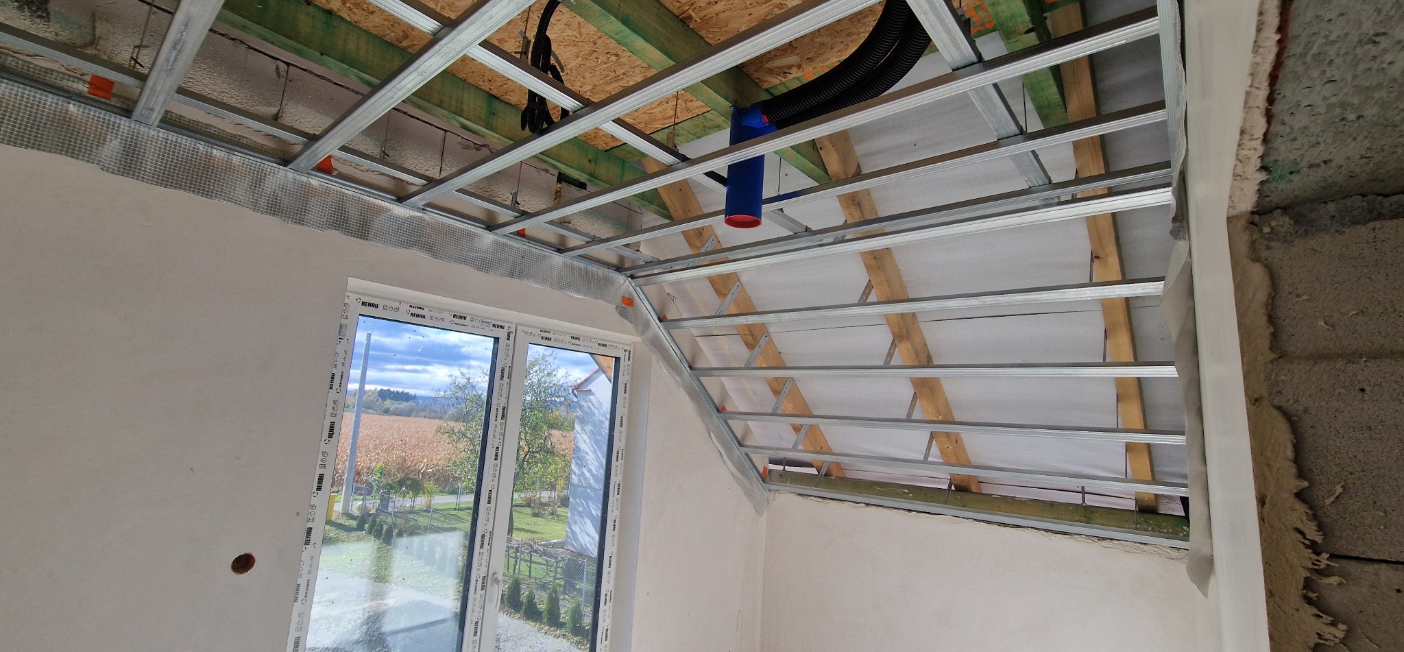 Skośny sufit poddasza z metalową konstrukcją pod płyty. Widoczne drewniane elementy dachu i instalacja wentylacyjna. Okno z widokiem na pole. Ściany przygotowane do wykończenia.