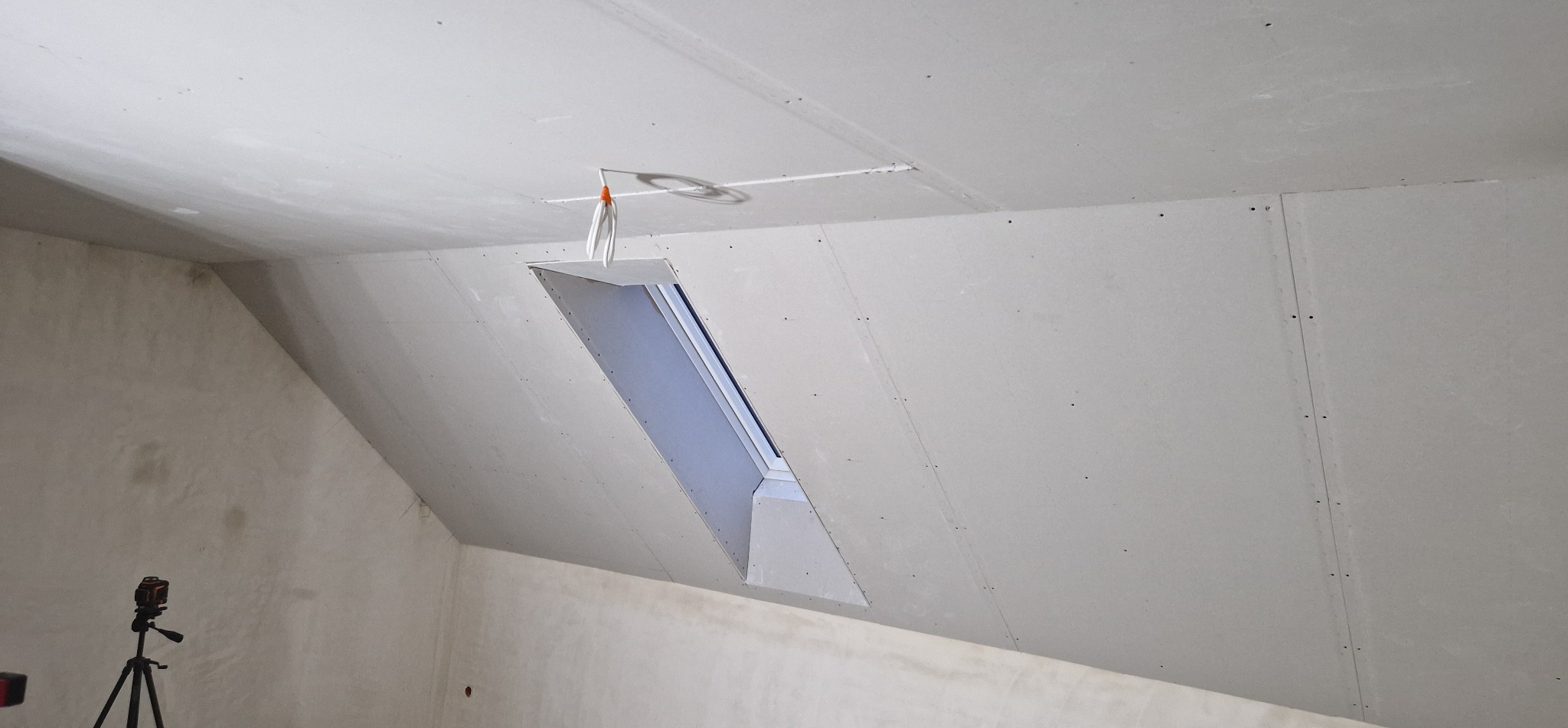 Poddasze z oknem dachowym po montażu płyt gipsowo-kartonowych, widoczne łączenia płyt i wystające przewody. W rogu pomieszczenia stoi laser krzyżowy na statywie.