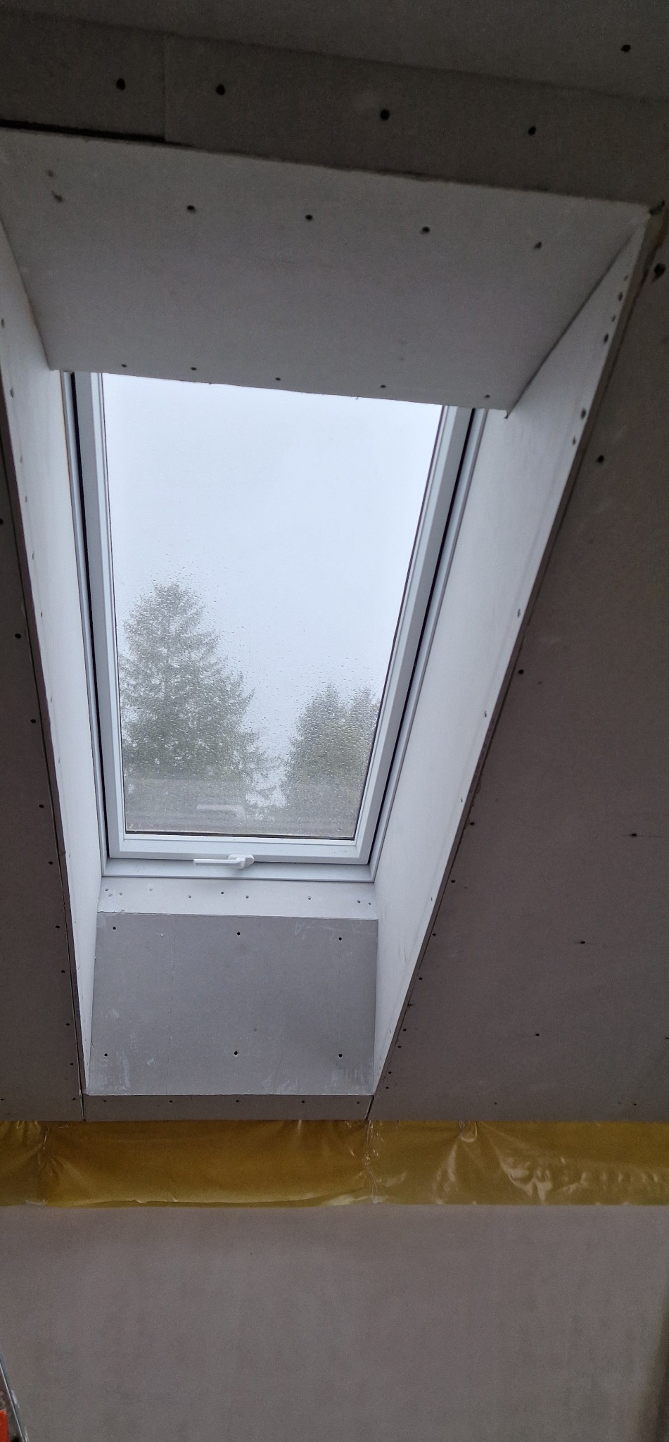 Montaż okna dachowego z obudową z płyty karton-gips, widok od dołu. Widoczne drzewa za oknem i żółta folia zabezpieczająca poniżej.