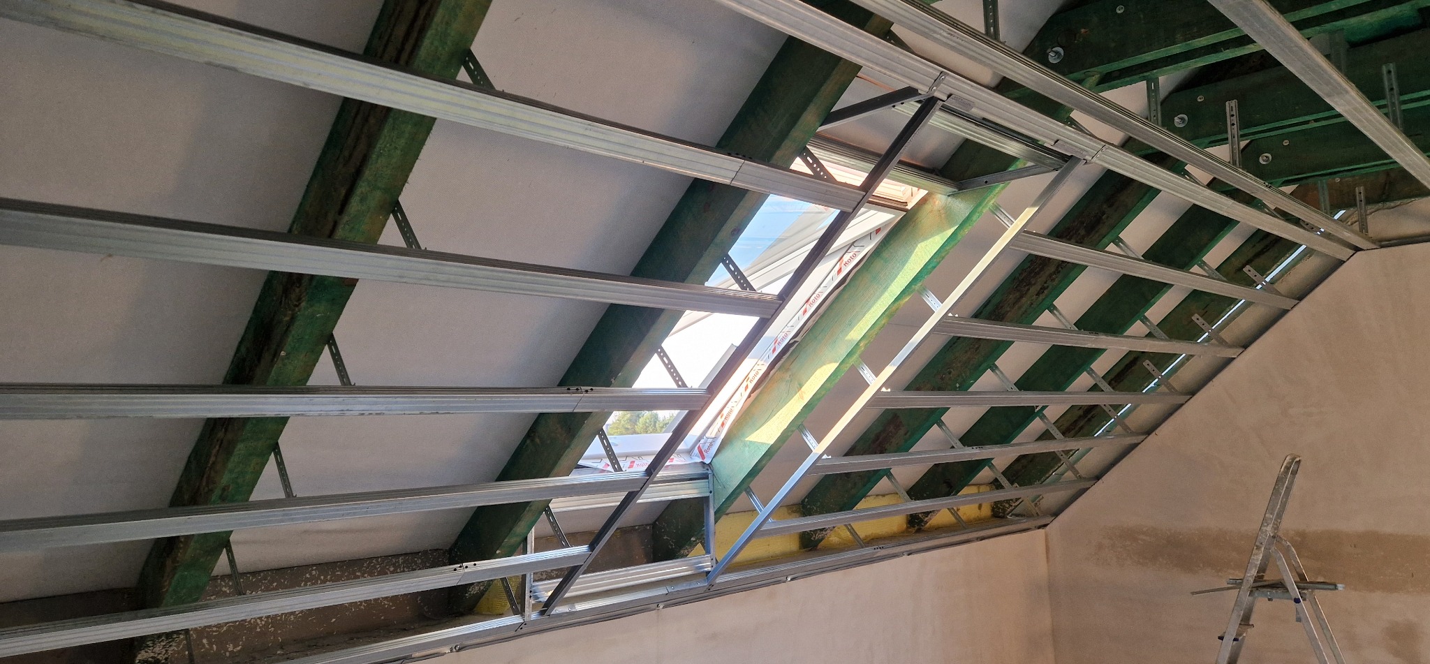 Szkielet sufitu podwieszanego z metalowych profili, widoczne drewniane elementy konstrukcyjne i okno dachowe. Prace wykończeniowe w poddaszu.