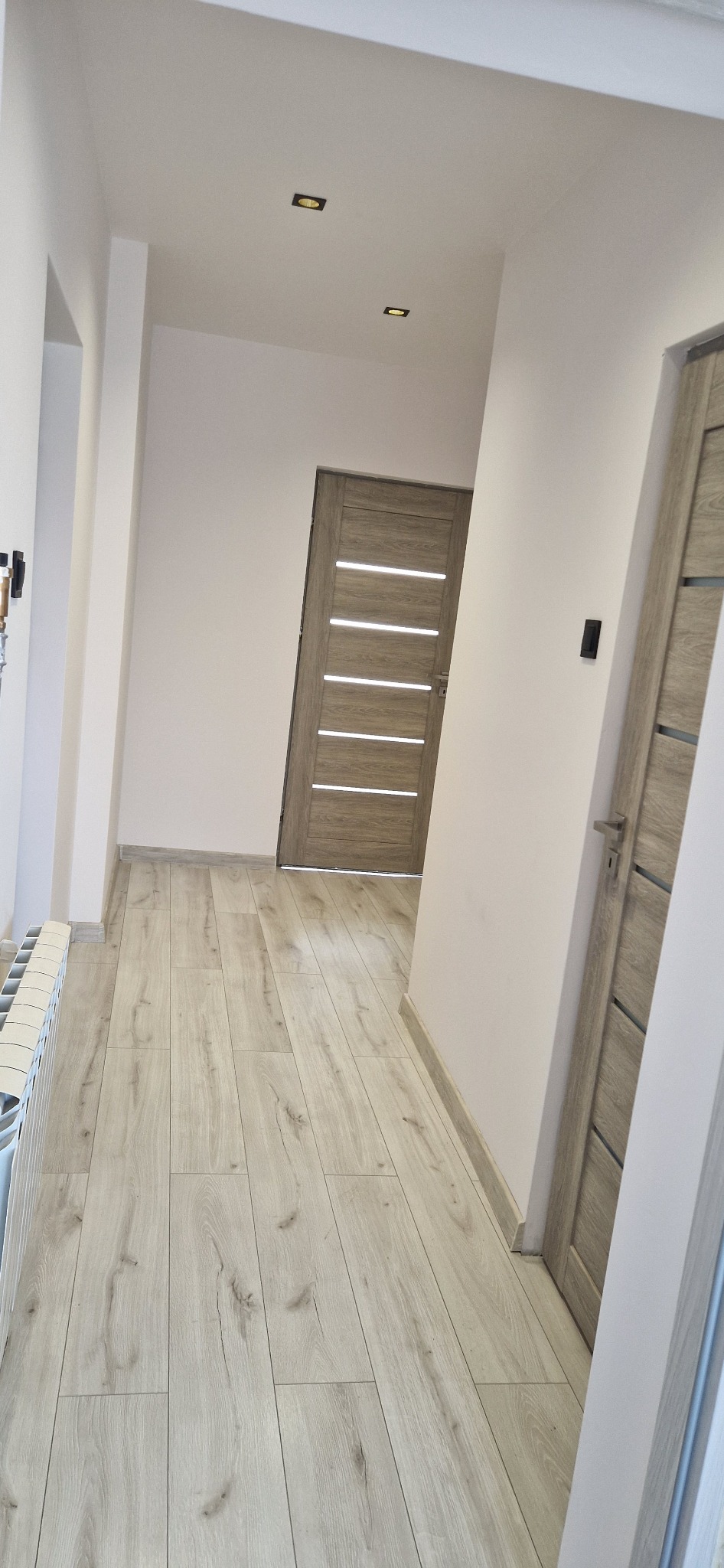 Jasny korytarz po remoncie: panele podłogowe, dwoje drzwi z poziomymi przeszkleniami, białe ściany i sufit z wbudowanym oświetleniem punktowym.