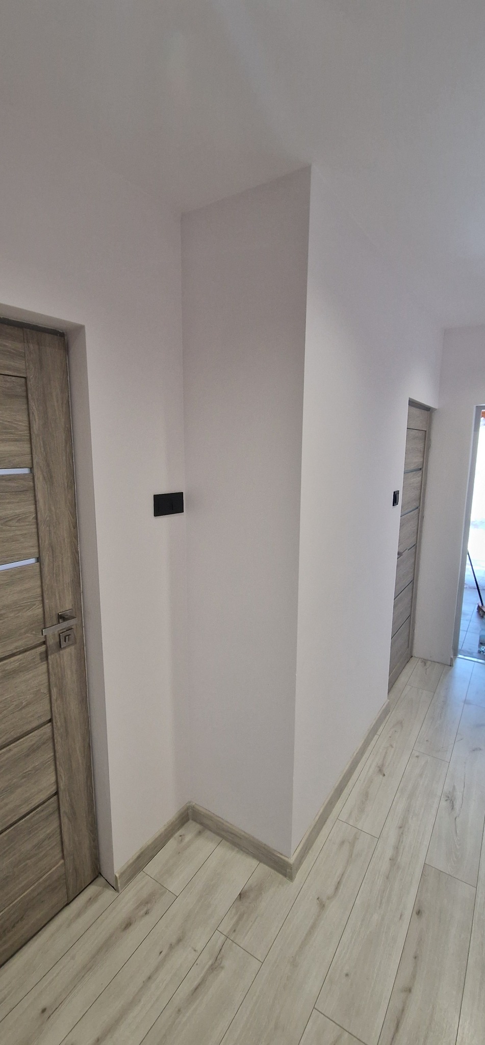 Wykończone wnętrze: gładkie, białe ściany, drewnopodobna podłoga, drzwi z drewnianą okleiną i czarne włączniki. Minimalistyczny design korytarza po remoncie.