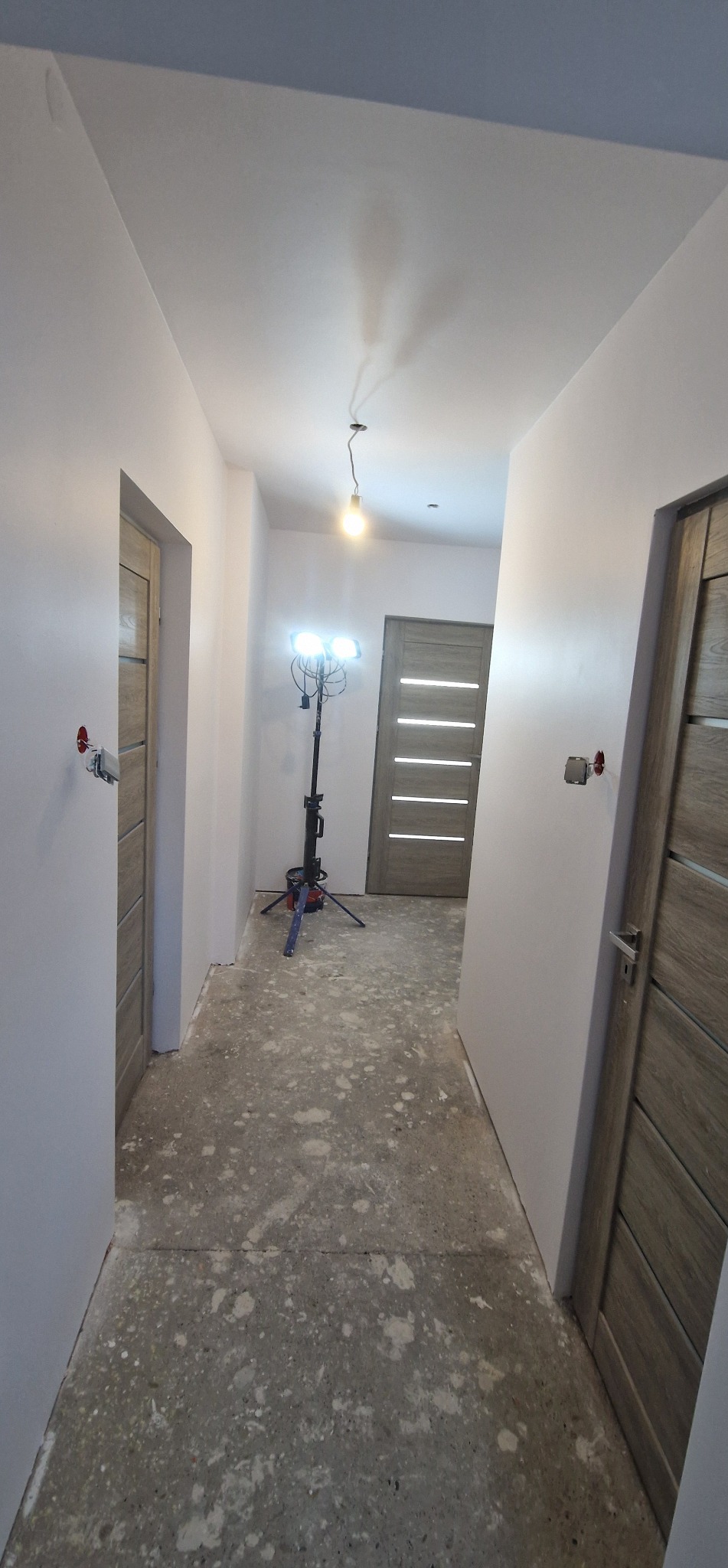 Korytarz w trakcie odświeżania ścian, widoczne dwie zamontowane drzwi, lampa budowlana i ślady po pracach na podłodze. Świeżo pomalowane ściany na biało.