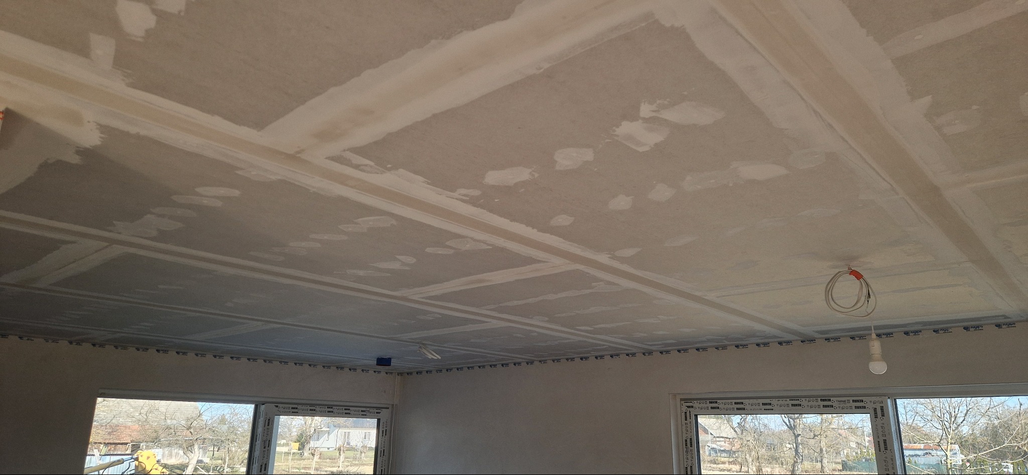 Sufit z płyt kartonowo-gipsowych, wstępnie wyszpachlowany, z widocznymi łączeniami i elementami instalacji elektrycznej. W tle okna z widokiem na zewnątrz.