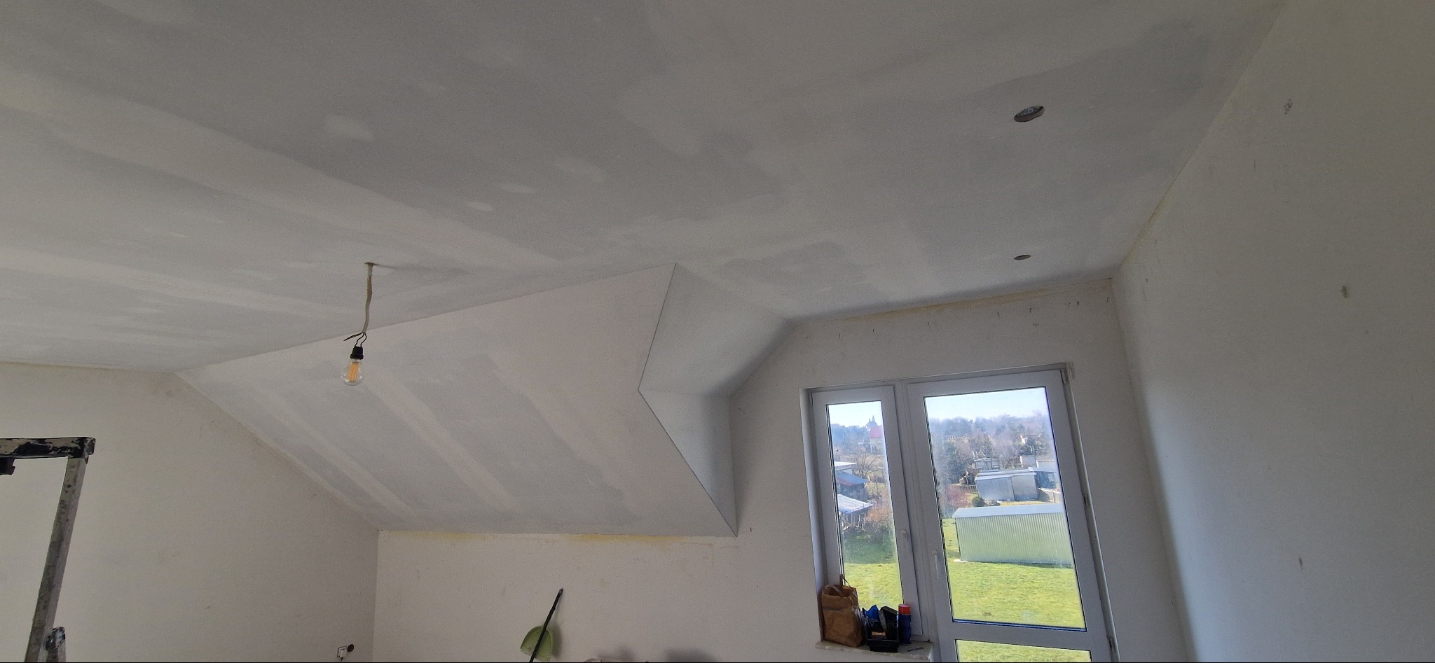 Poddasze po szpachlowaniu, widoczne łączenia płyt gipsowych na skosach i suficie, przygotowane do dalszych prac wykończeniowych. Okno z widokiem na zewnątrz.