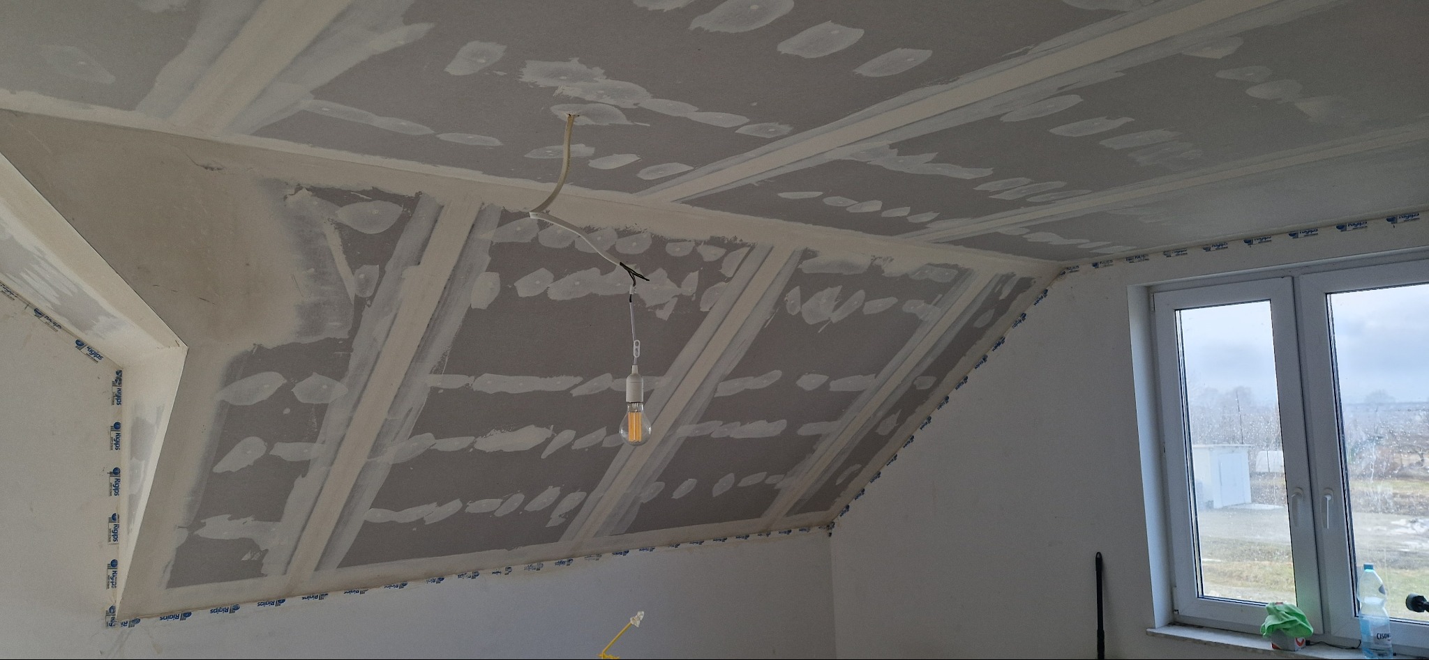 Skośny sufit z płyt kartonowo-gipsowych w trakcie prac wykończeniowych, widok z dołu, Radymno. Widoczne łączenia płyt i lampa wisząca.