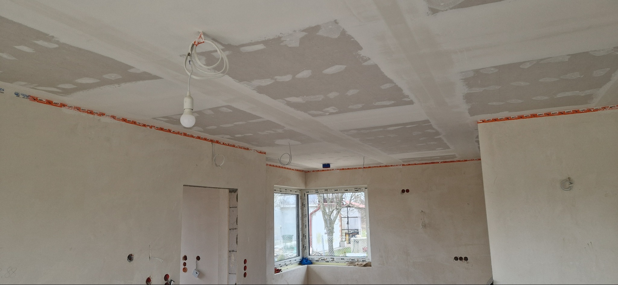 Wnętrze w trakcie remontu: ściany i sufit po szpachlowaniu, z widocznymi przewodami elektrycznymi i oknem narożnym. Etap przygotowania do malowania.
