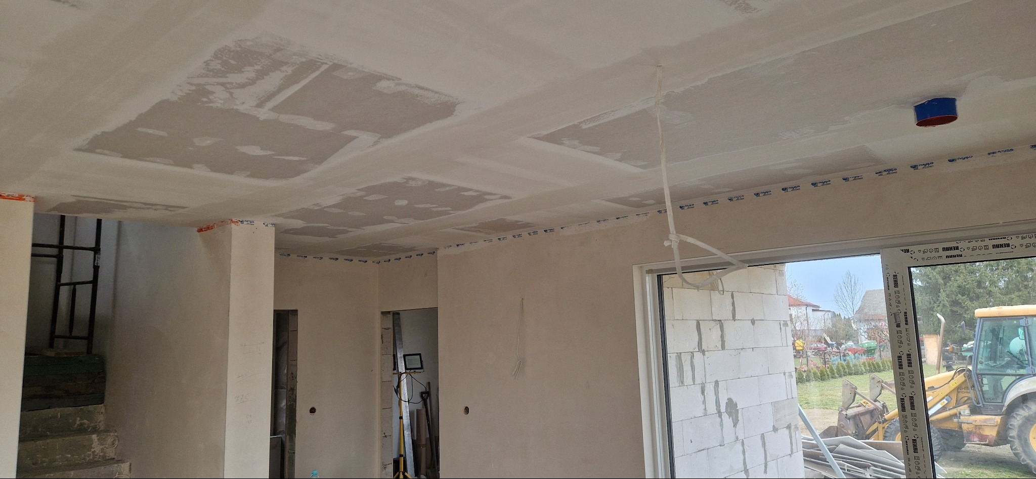 Wnętrze w trakcie prac wykończeniowych: sufit z widocznymi łatami po szpachlowaniu, ściany przygotowane do dalszych prac. Widoczne okno i fragment traktora na zewnątrz.
