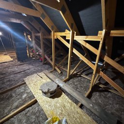 Poddasze w trakcie prac izolacyjnych: wełna mineralna między krokwiami, widoczne elementy konstrukcji drewnianej i rolka sznurka. Ujęcie z dołu, ciemne tło.