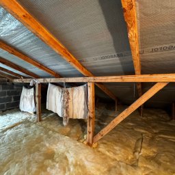 Poddasze z widoczną izolacją z wełny mineralnej, drewnianą konstrukcją więźby dachowej i folią paroizolacyjną. Część poddasza zasłonięta białym materiałem.