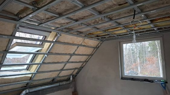 Skośny sufit z oknami połaciowymi w trakcie ocieplania wełną mineralną i montażu stelaża w Skierniewicach. Widoczna instalacja oświetleniowa.