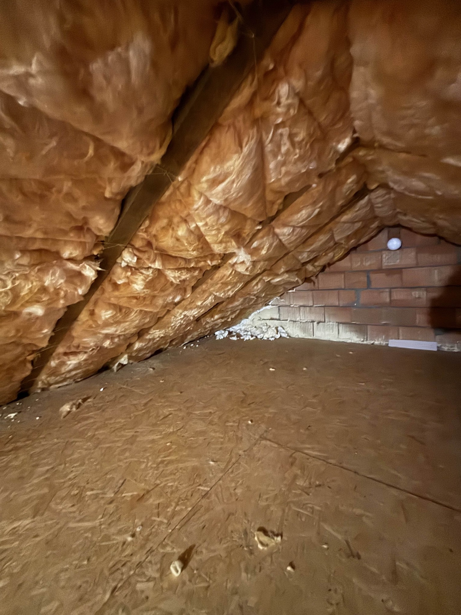 Ocieplone wełną mineralną skośne poddasze z drewnianą konstrukcją i fragmentem ściany z czerwonej cegły w tle. Widoczne niedociągnięcia w izolacji.